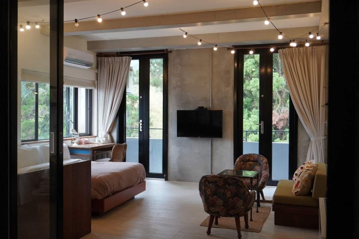 A modern bedroom with a bed, a desk, and a TV. There are curtains, a window, and a balcony with a view.
