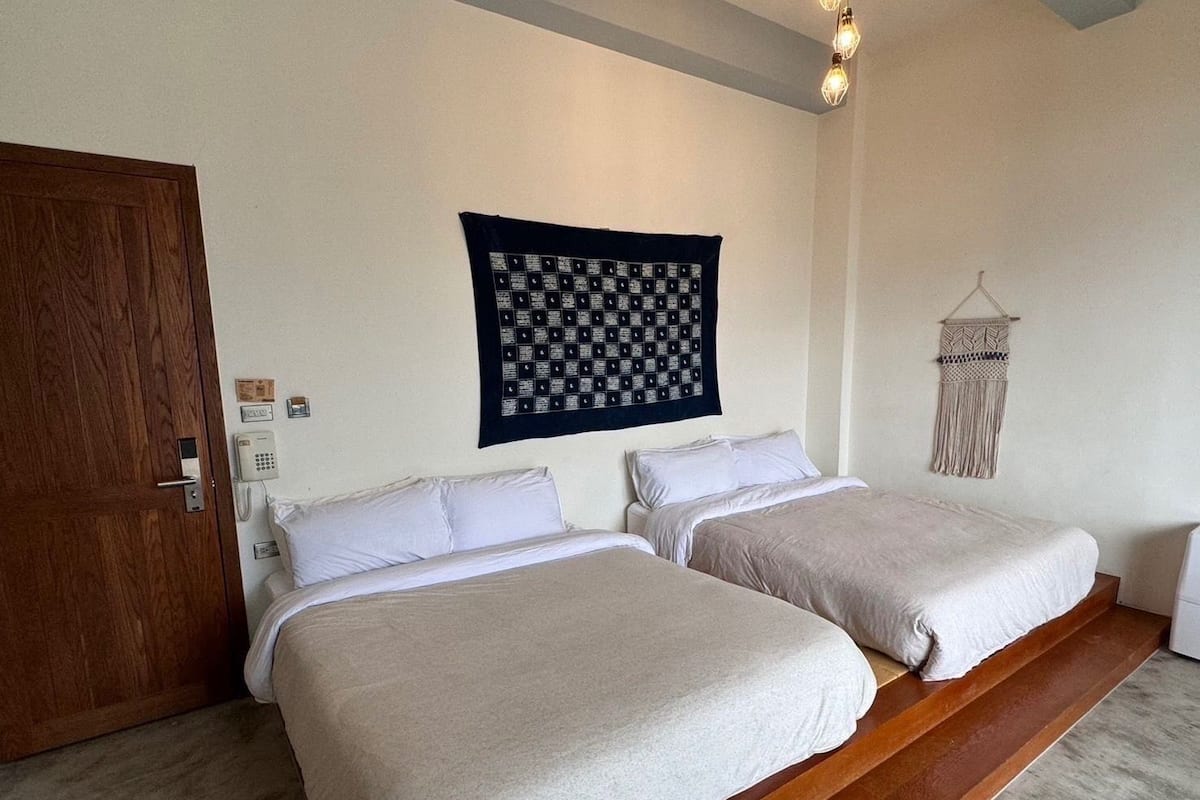 A bedroom with two beds, a wooden door, a wall-mounted phone, and a hanging macrame wall hanging.