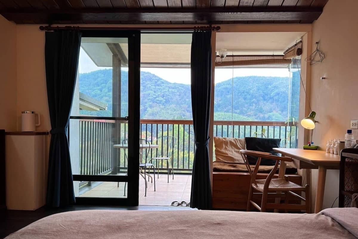 A room with a bed, a desk, and a balcony with a view of mountains.