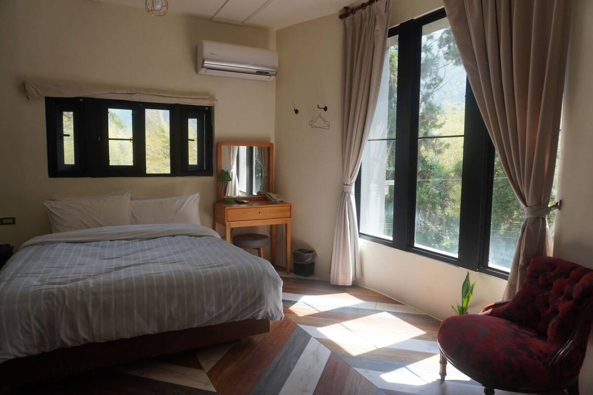 A bedroom with a bed, a nightstand, a chair, a window with curtains, and an air conditioning unit.