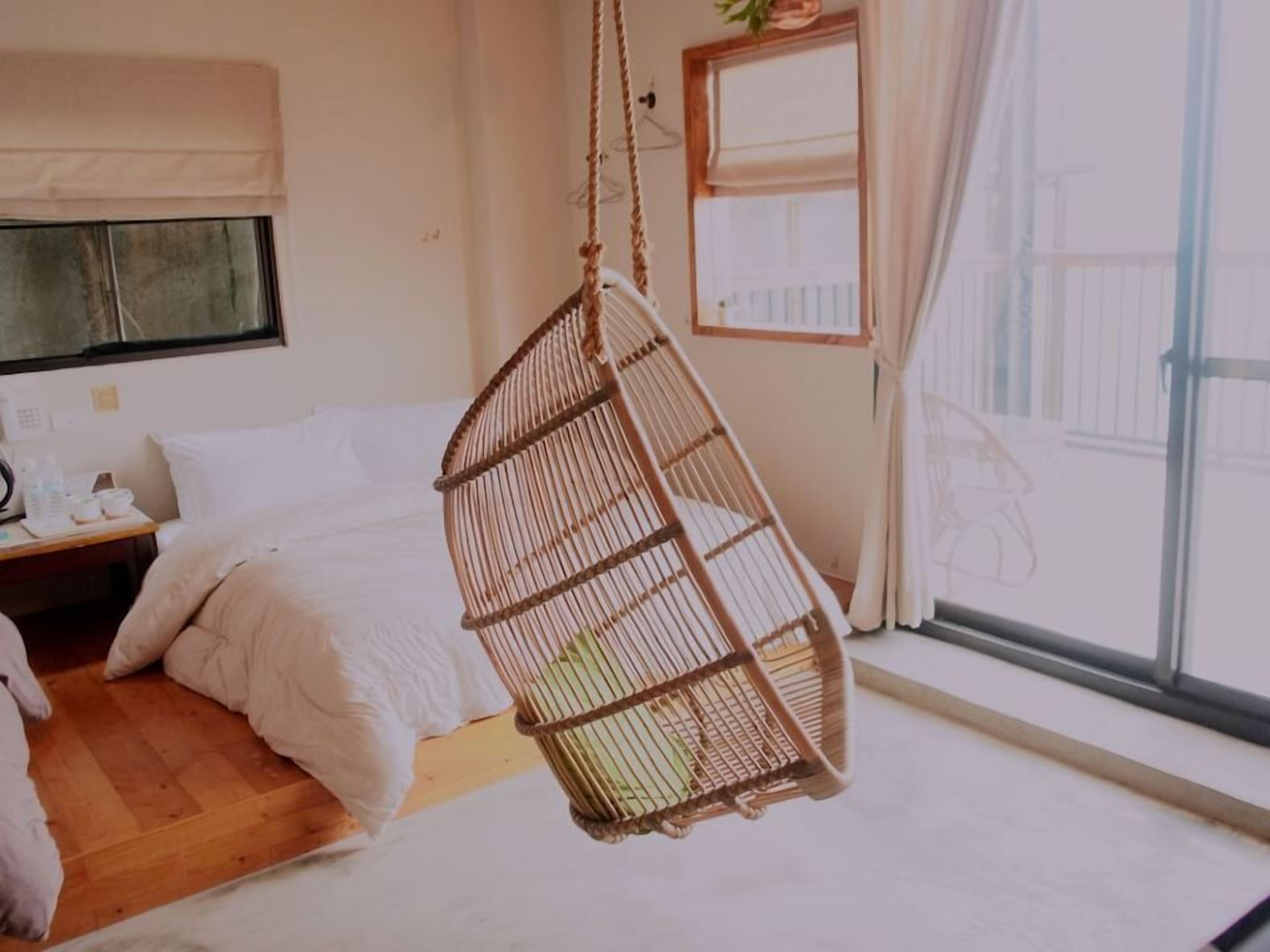 A bedroom with a hanging chair, a bed, a bedside table with toiletries, and a window with curtains.