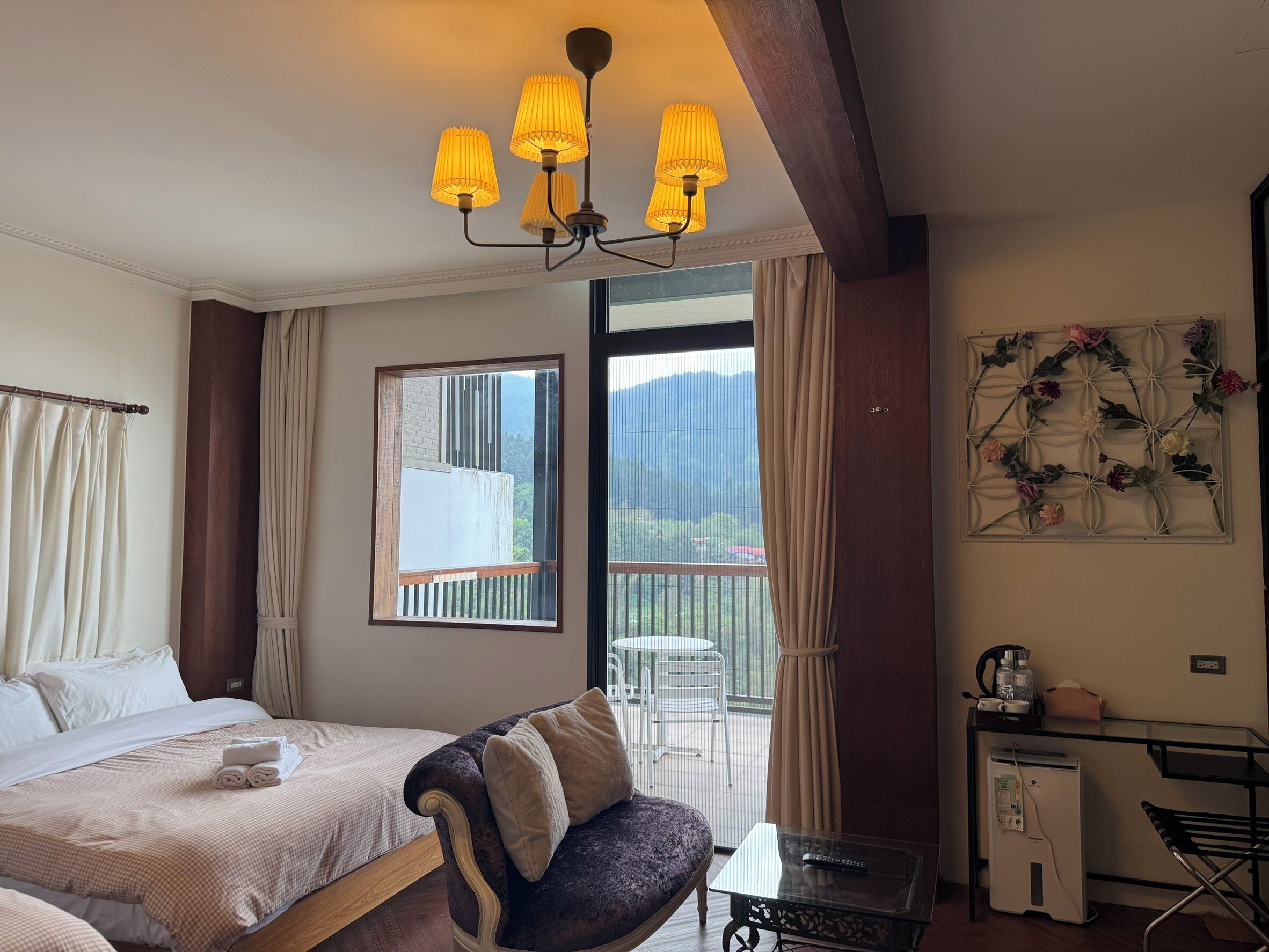 A hotel room with a bed, a sofa, a balcony with a table and chairs, and a desk with a refrigerator.
