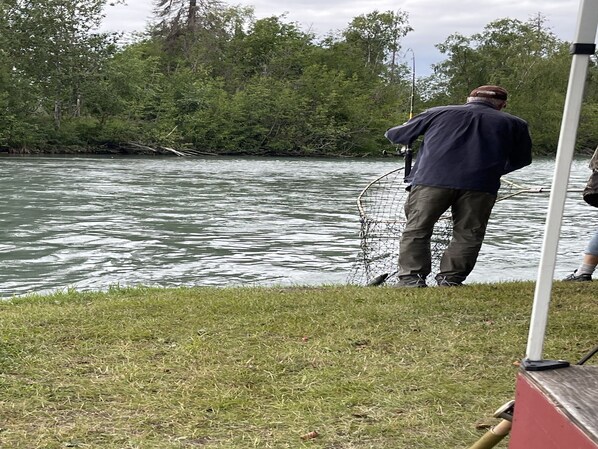 Una persona de pie junto a un río con una caña de pescar, rodeada de vegetación.