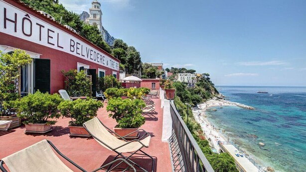 Un hotel con fachada roja, terraza al aire libre y vista al mar.