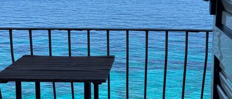 A balcony with a table and chair overlooking a turquoise sea and distant mountains.