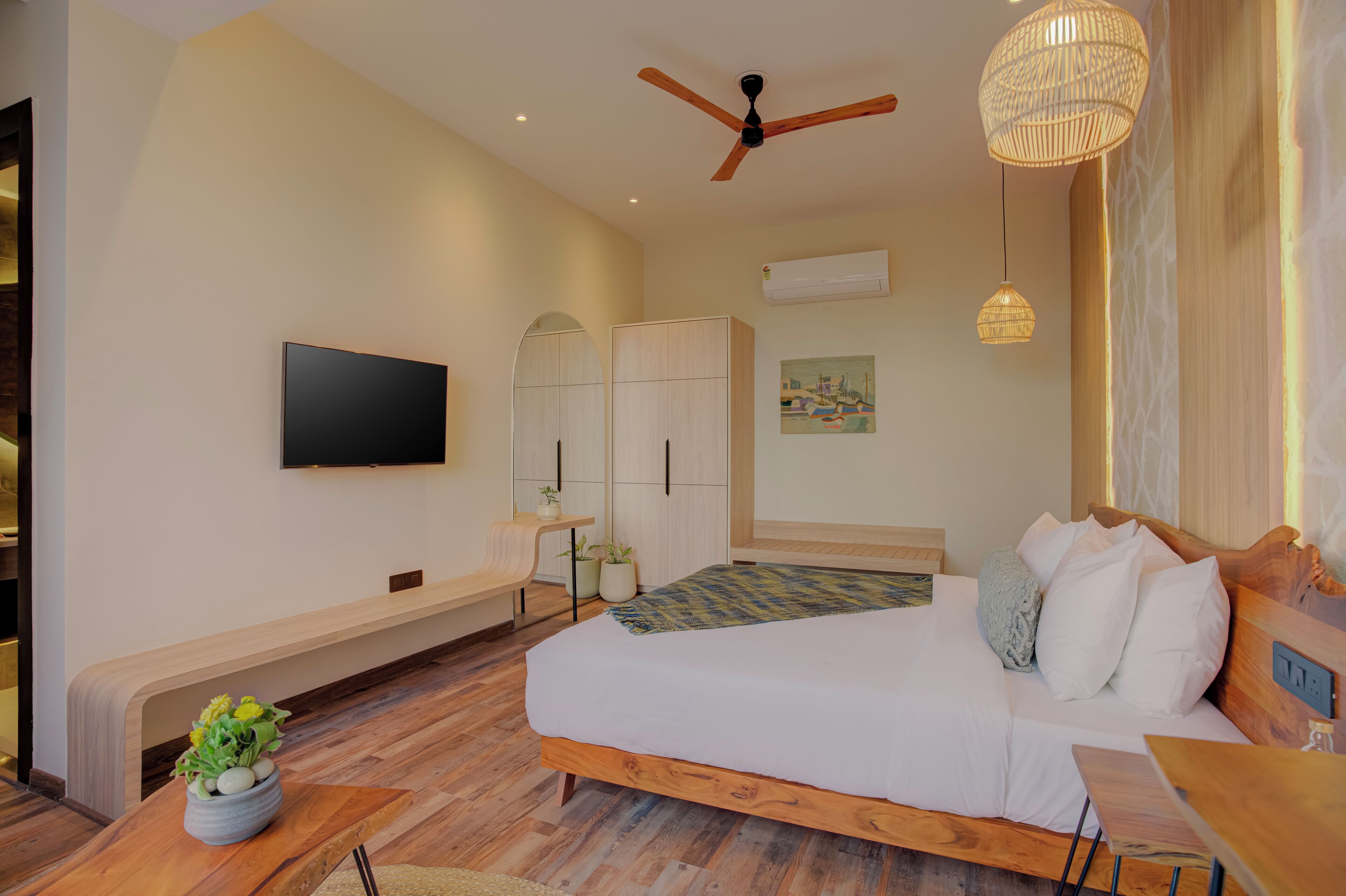 Une chambre moderne avec un lit en bois, un ventilateur de plafond, un téléviseur fixé au mur et un petit coin salon agrémenté d’une plante.