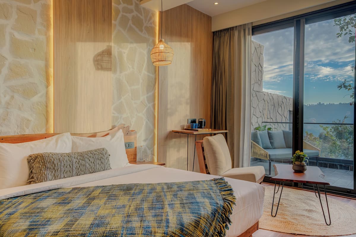 A bedroom with a large bed, a bedside table with a lamp, a view of the outdoors through a large window, and a small table with a plant.