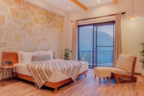 A bedroom with a stone wall, a bed with a wooden headboard, a bedside table, a chair, and a balcony with a mountain view. - Moshams - A Home with Nature & Elegance, near Rishikesh (Narendranagar)