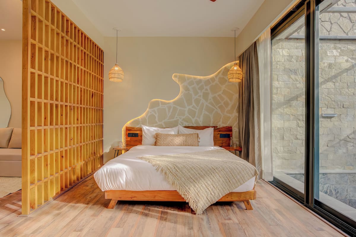 A modern bedroom with a wooden headboard, a large bed, and a view of stone walls through a glass door.