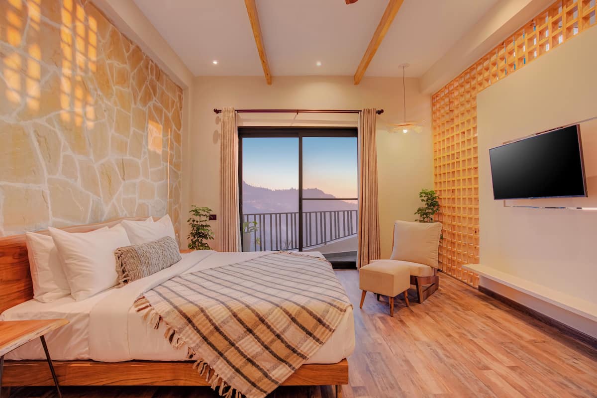 A bedroom with a large bed, a TV mounted on the wall, a balcony with a view, and a wooden floor.