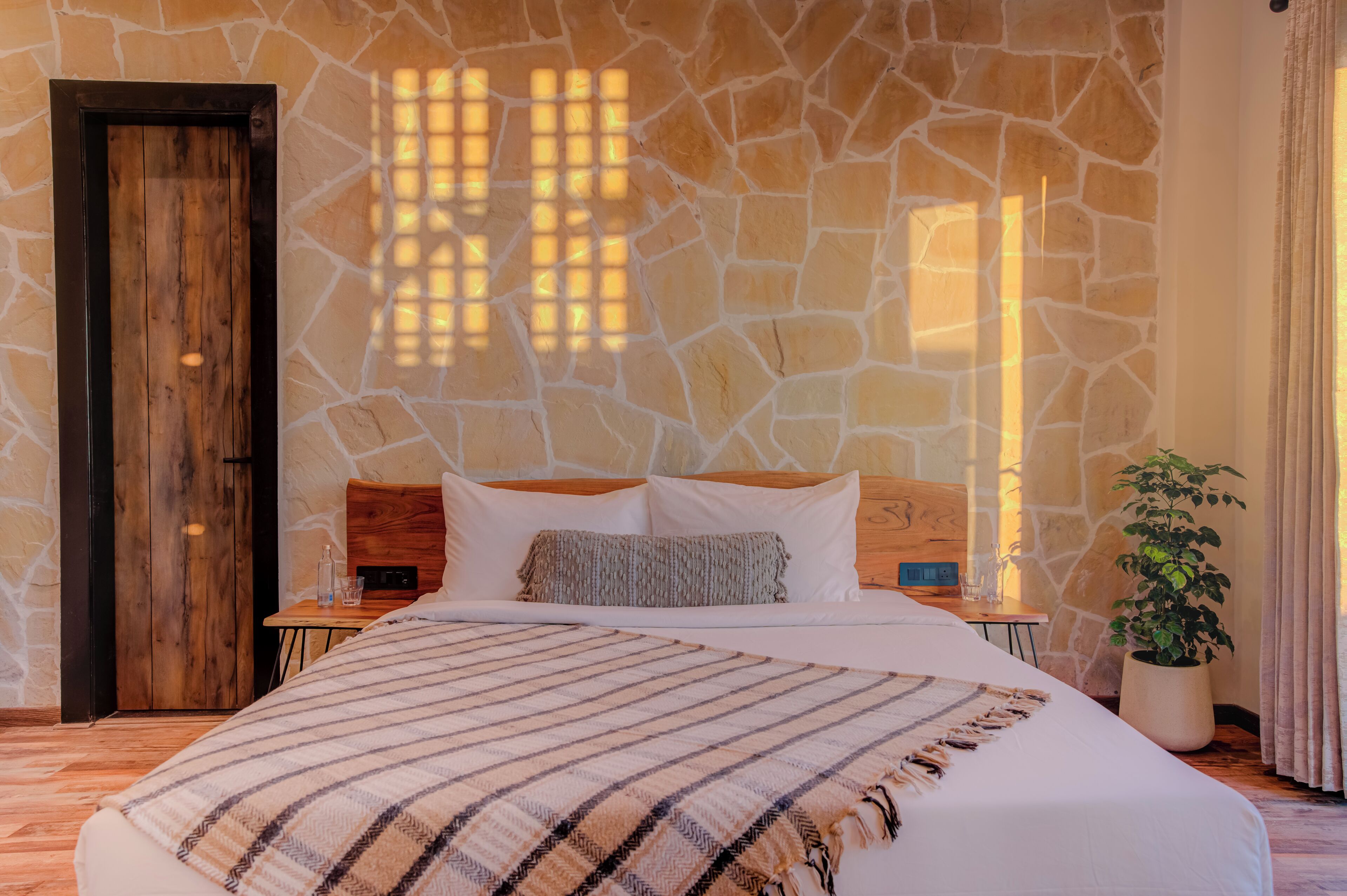 Une chambre avec un mur en pierre, une porte en bois, un lit recouvert d’une couverture à rayures, une table de chevet avec une lampe et une plante en pot.