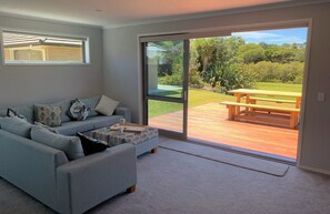 A modern living room with a grey sofa, a coffee table, and a sliding glass door leading to an outdoor patio with a wooden deck and picnic table. - Riverfront gem in Ruakaka (Ruakākā)