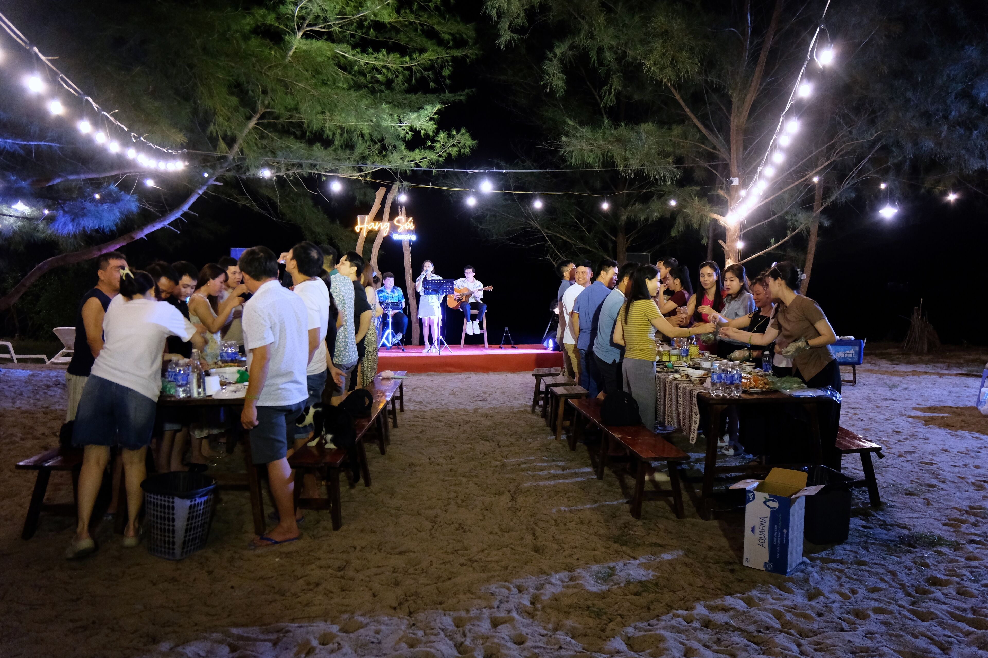 An outdoor event with people gathered around tables, string lights overhead, and a stage in the background.