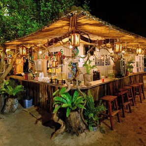 Eine rustikale Außenbar mit Holzelementen, hängenden Lichtern und viel Grün.