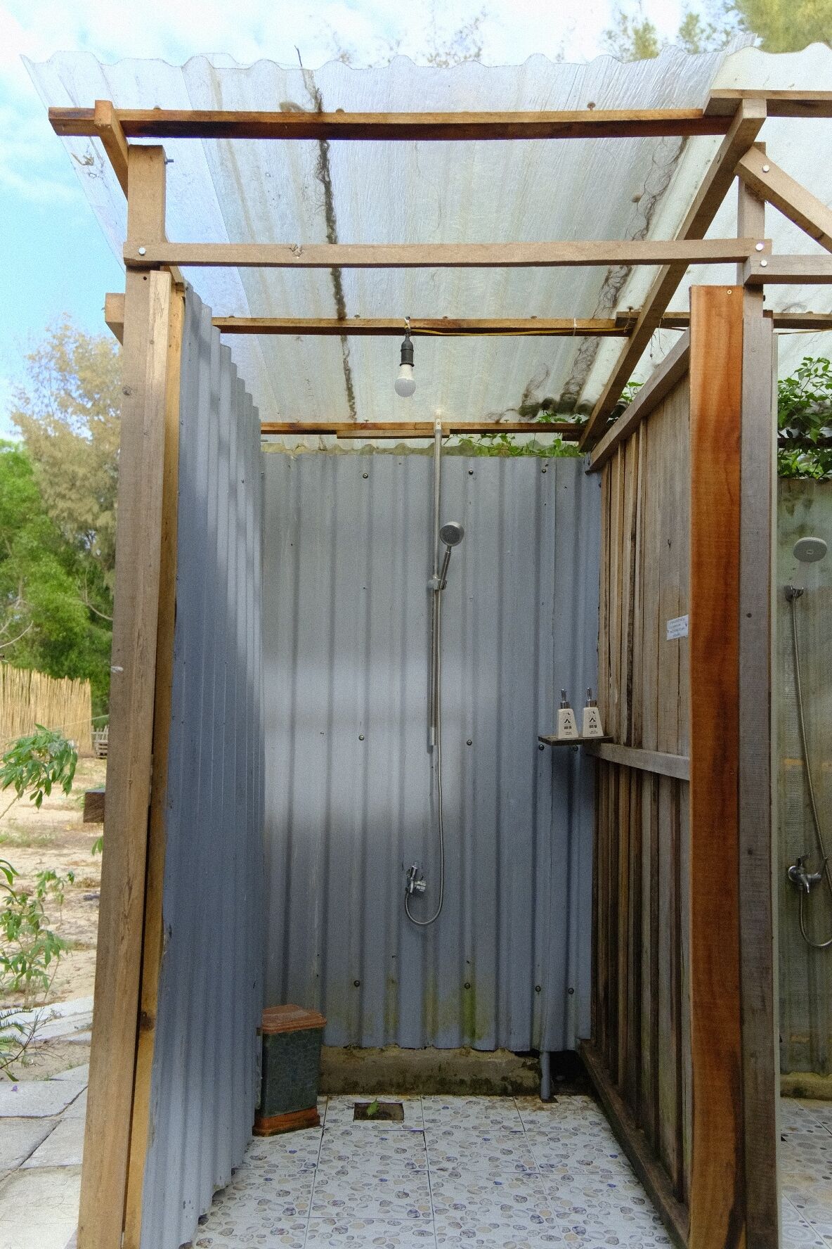 A small, enclosed outdoor shower area with a metal roof and wooden frame, featuring a showerhead and a handwashing station.