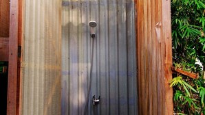 A small, enclosed outdoor shower area with a corrugated metal wall, a wooden roof, and a metal barrel.