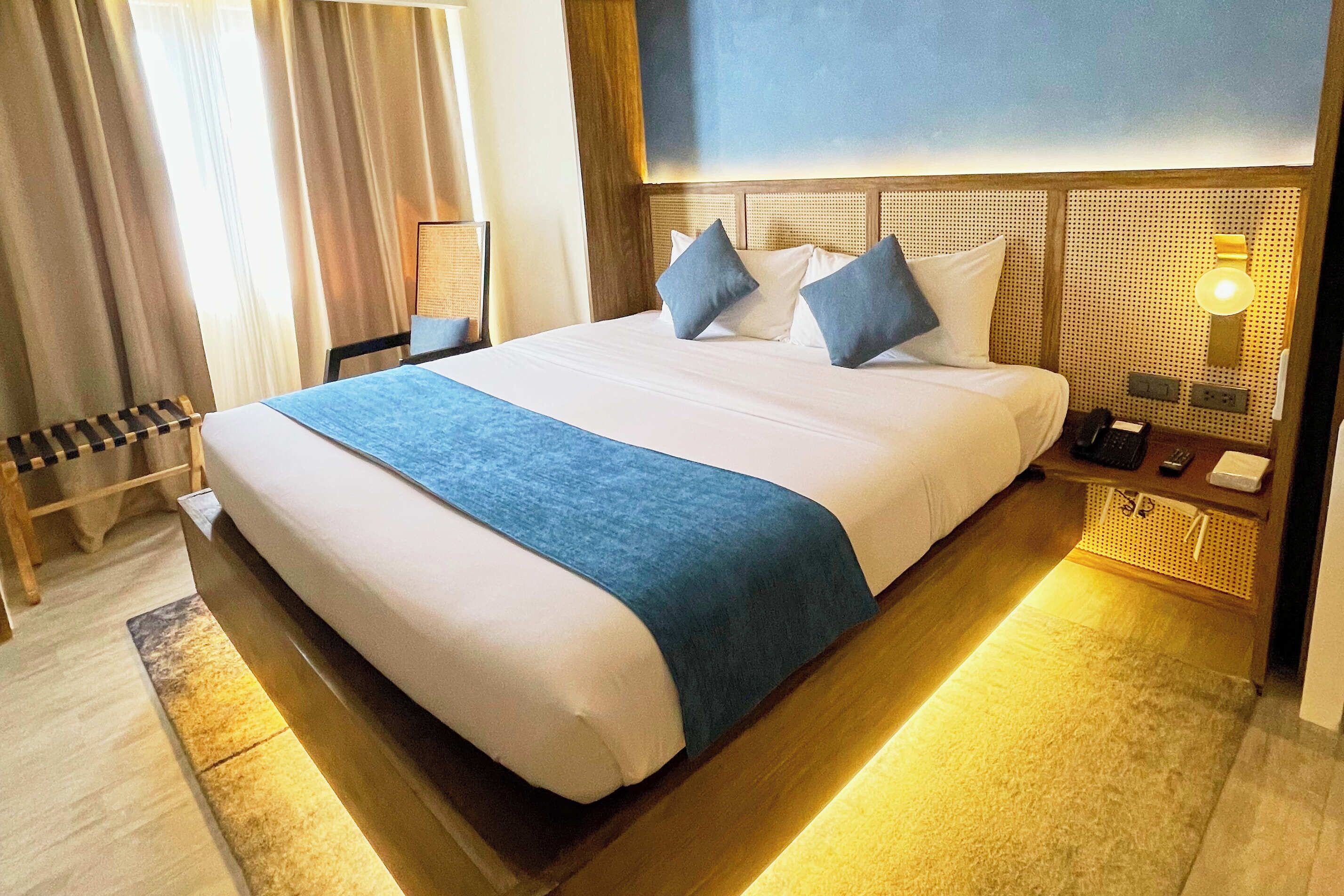 A hotel room with a large bed, a bedside table with a lamp, and a view of the outdoors through the window.