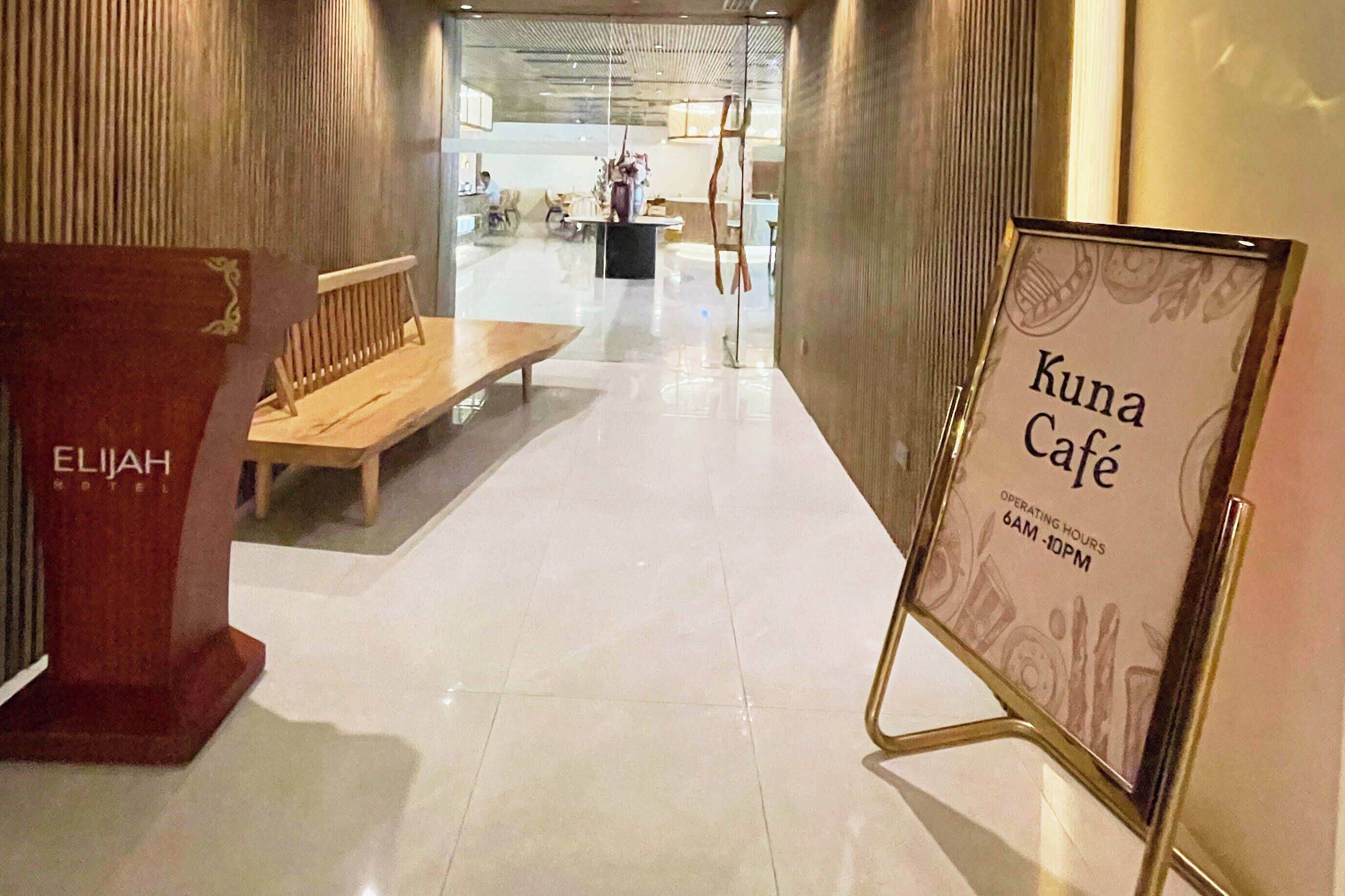 A hallway with a wooden bench, a sign for 'Kuna Café' with operating hours, and a 'ELIJAH HOTEL' sign.