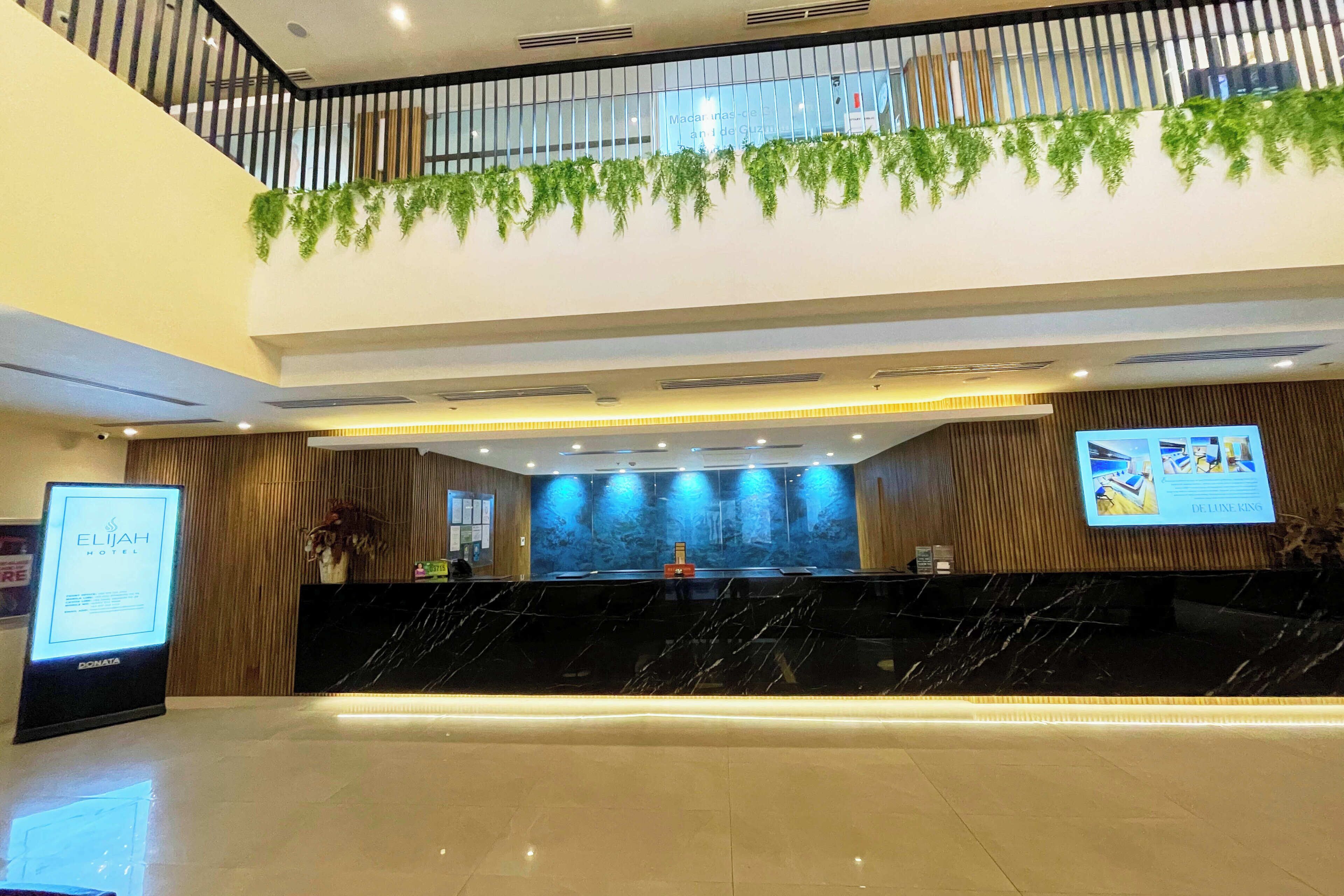 A hotel reception area with a black marble counter, a digital screen displaying hotel information, and a balcony with greenery.