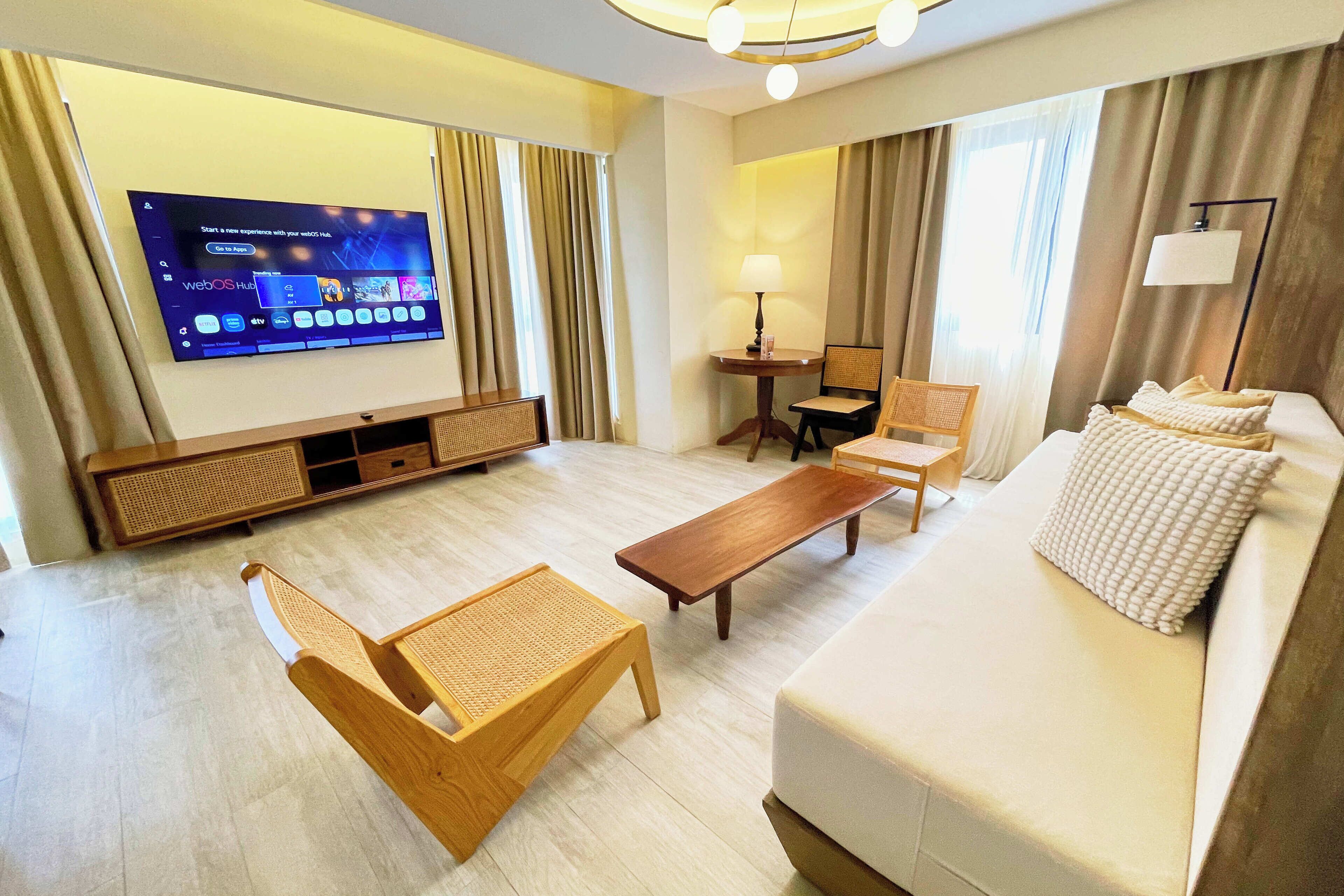 Ein Hotelzimmer mit Flachbildfernseher an der Wand, einer Couch, einer Holzbank, einem kleinen Tisch und einer Lampe.