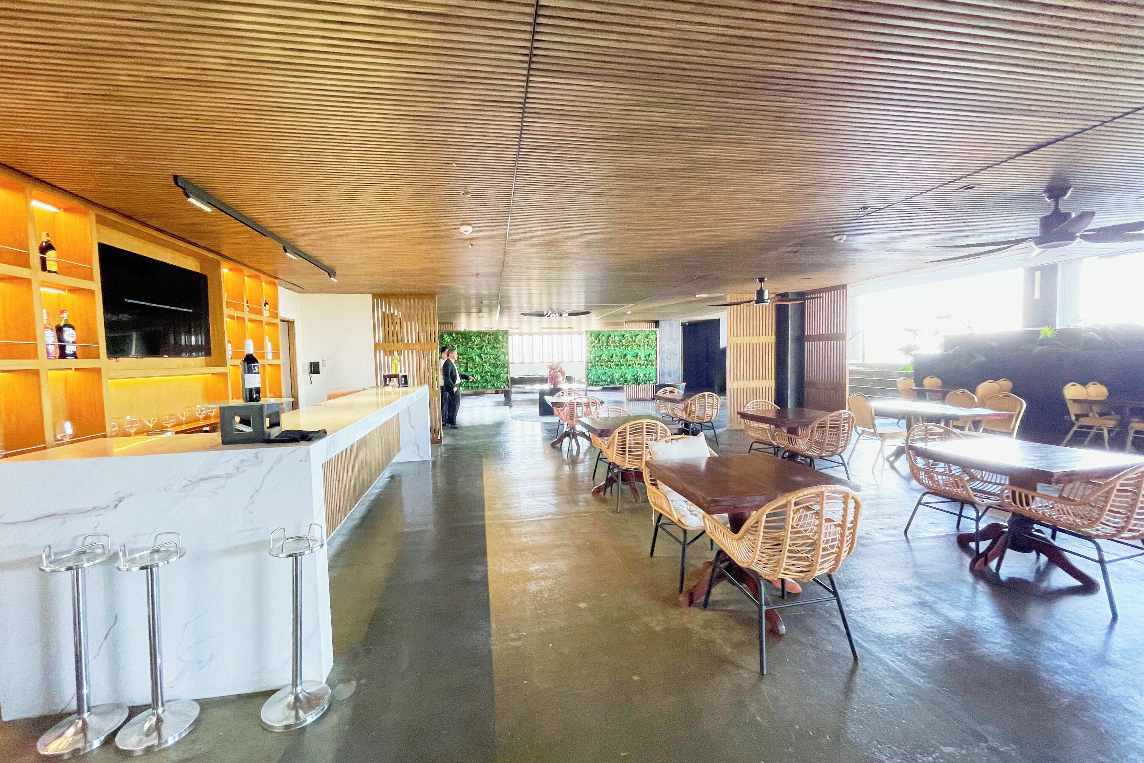 A spacious indoor area with a wooden ceiling, a bar area with shelves of bottles, wicker chairs, and tables.