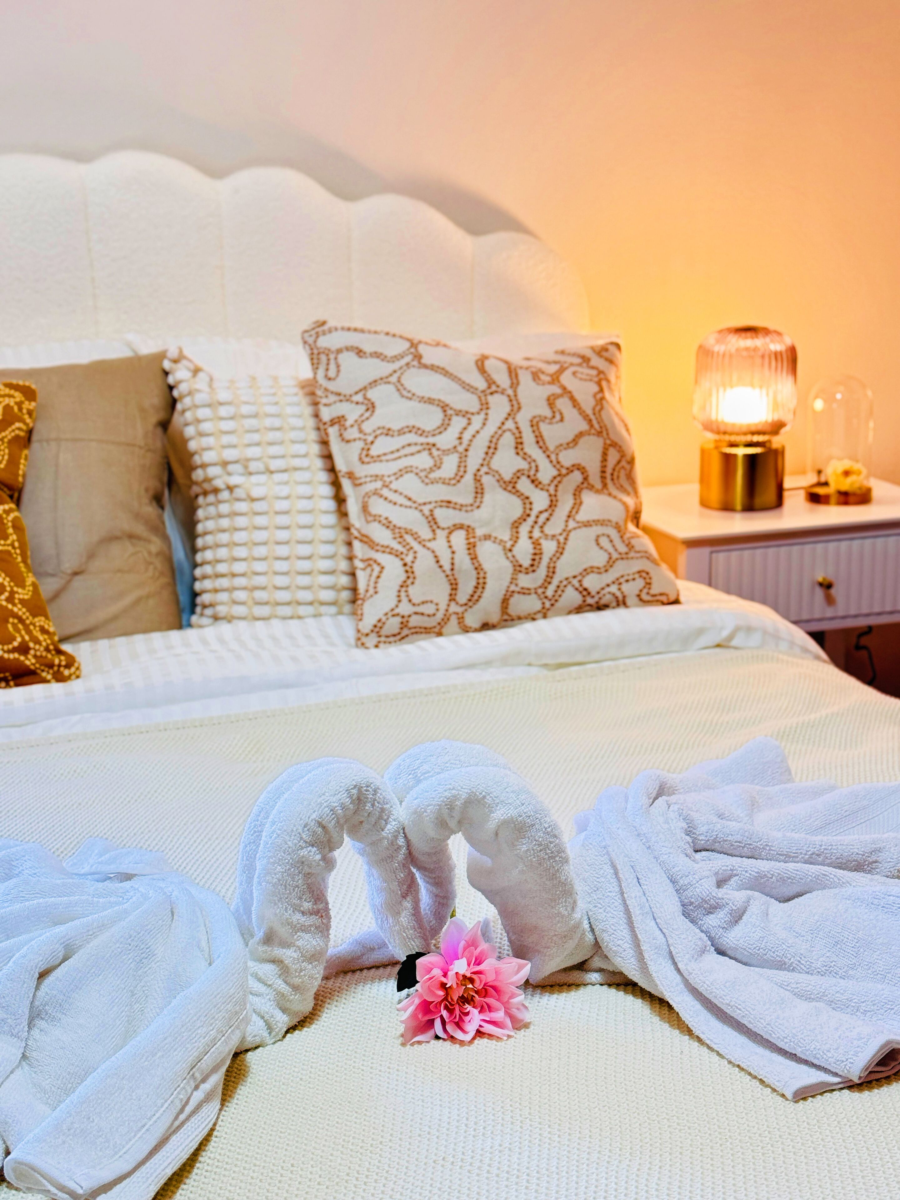 A neatly made bed with white and patterned pillows, a decorative lamp, and towels arranged on the bed.