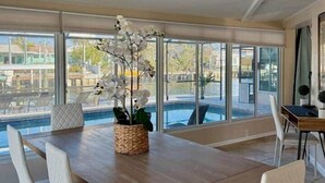 A dining area with a wooden table and white chairs, a woven basket with orchids, and large windows overlooking a pool. - Luxury Oceanfront 3BR with Pool, Dock & Boat Lift (Cortez)