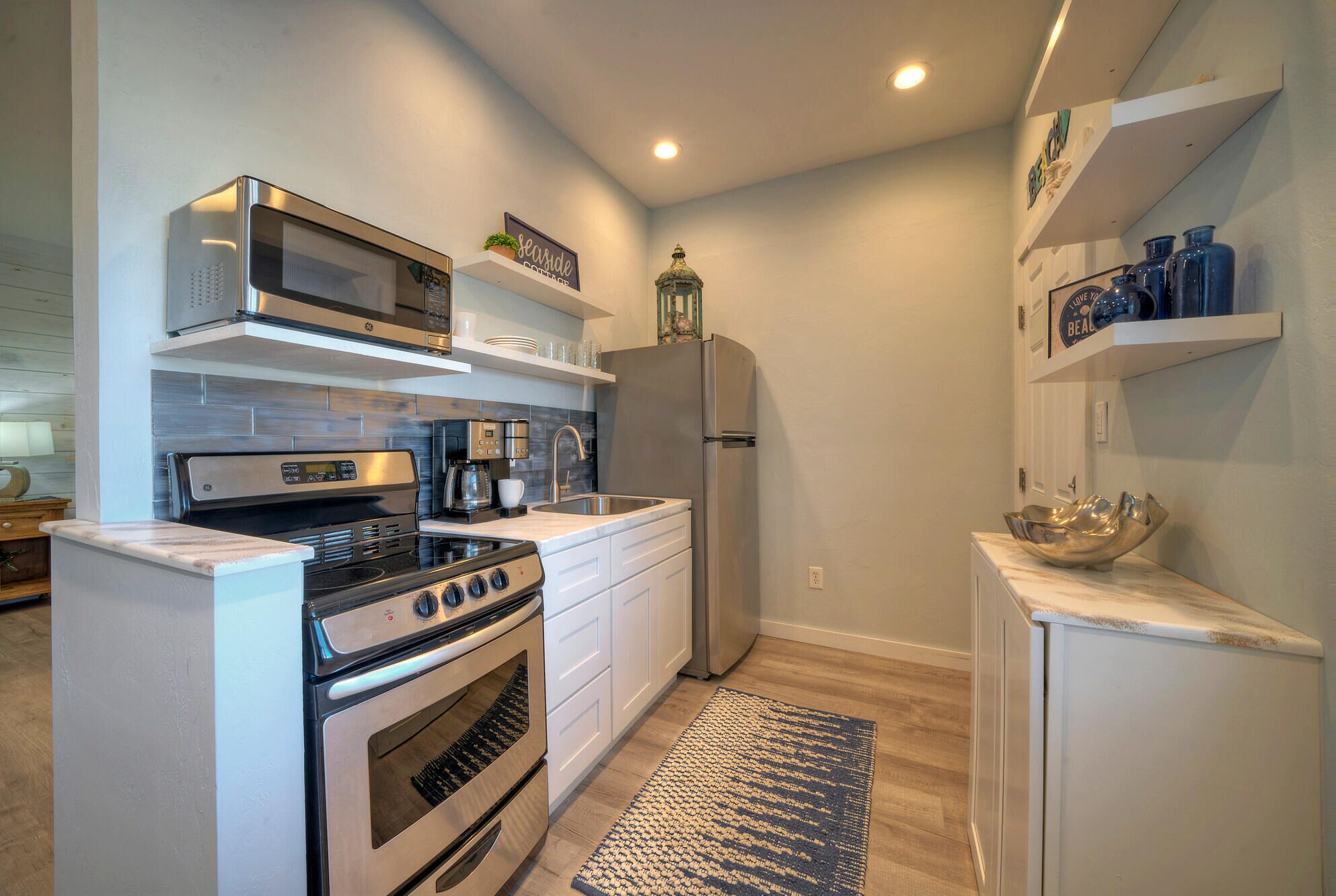 Cocina compacta con estufa, horno, microondas y refrigerador. Cuenta con gabinetes blancos, una cafetera y un tapete en el suelo.