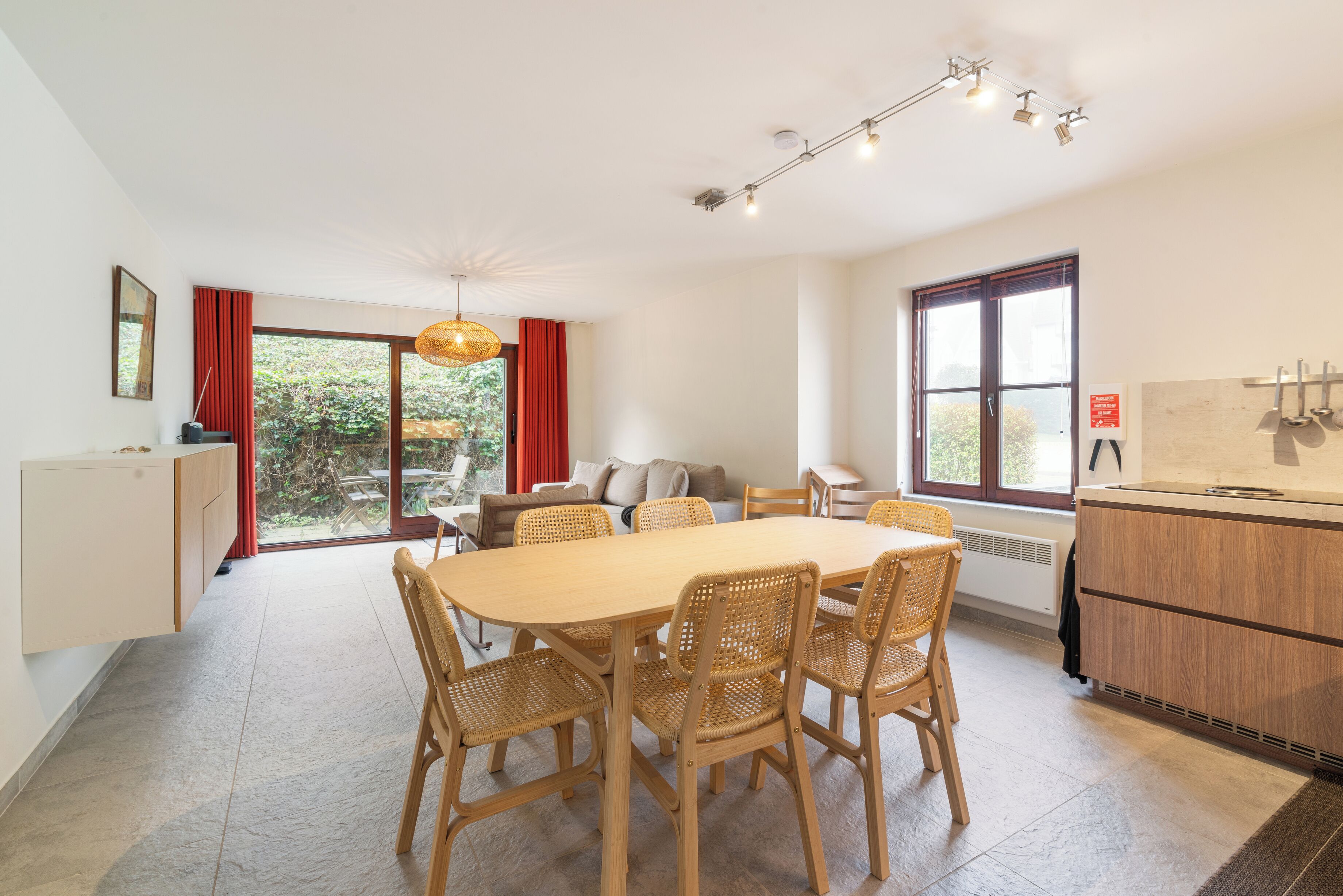 A modern dining area with a large wooden table and wicker chairs, a sofa, and a kitchen area with a stove.