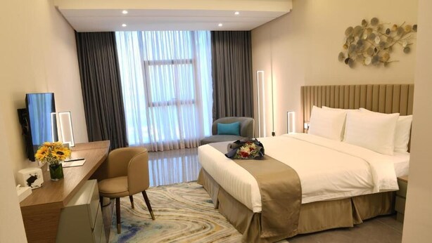 A hotel room with a bed, a desk, a chair, a large window with curtains, and a decorative wall piece. - The Mark Hotel  (Manama)