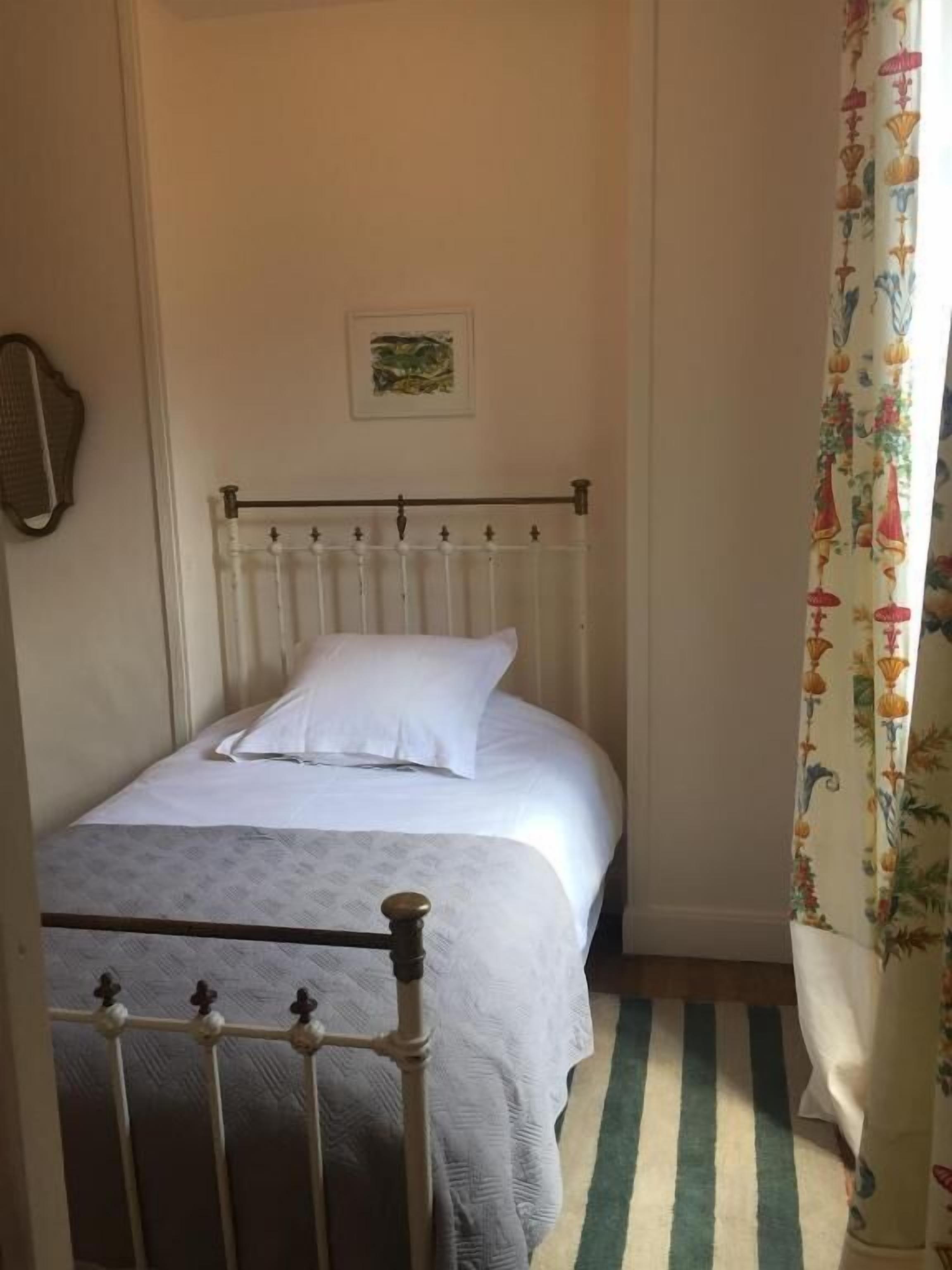 Una cama individual con cabecera y piecera blancos, una pared lisa con un pequeño cuadro enmarcado, y una alfombra a rayas en el suelo.