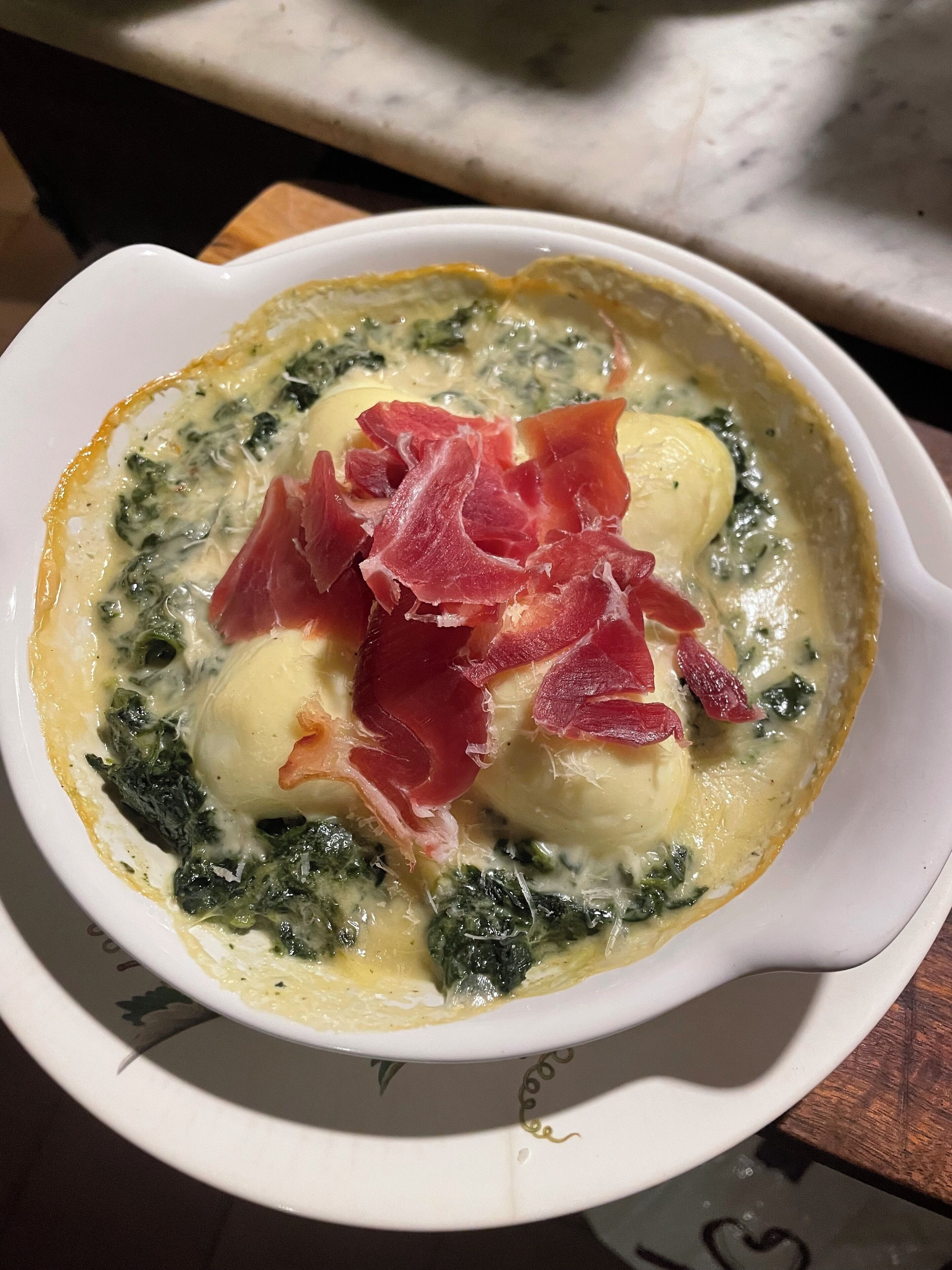 A white ceramic dish with a heart-shaped handle containing a creamy soup with spinach and pieces of prosciutto.