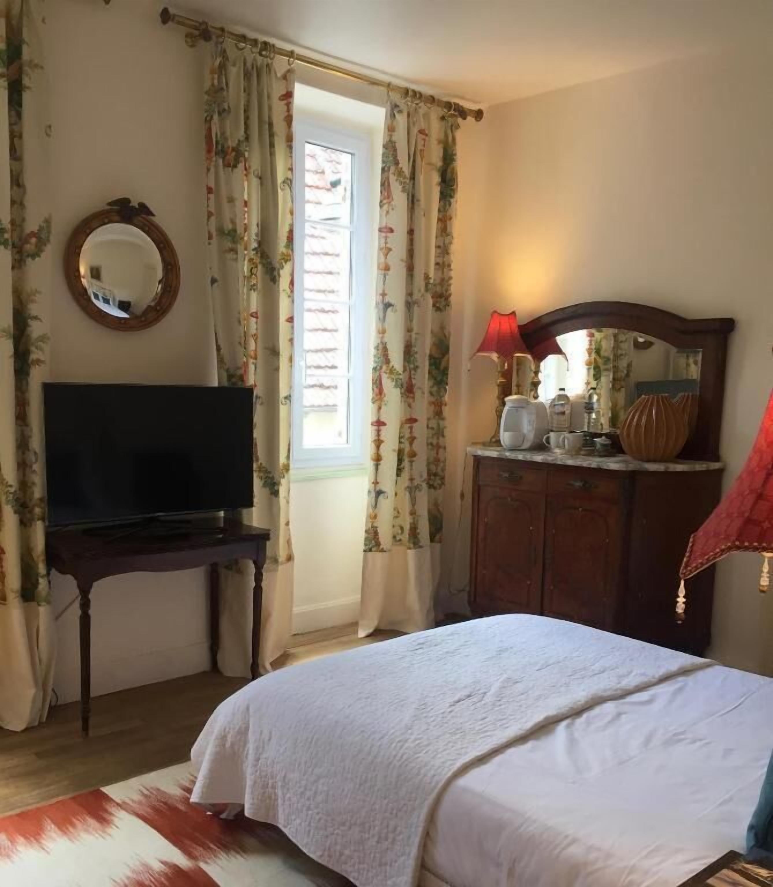 A bedroom with a bed, a TV on a stand, a dresser, a mirror, and curtains.