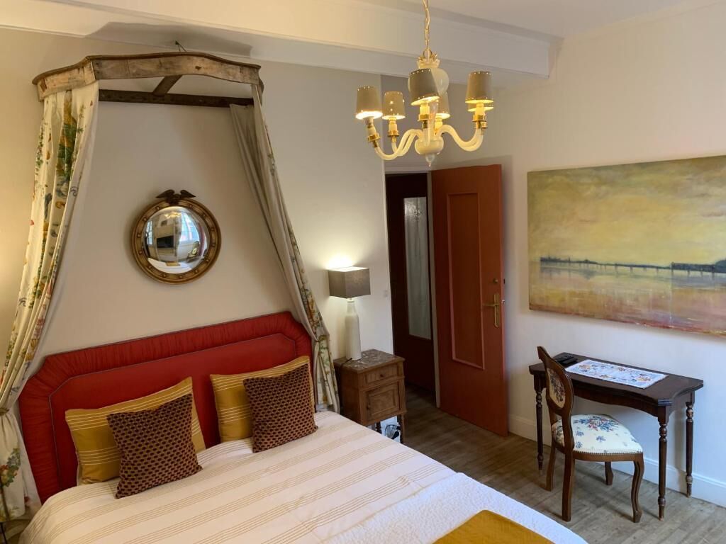 Un dormitorio con cabecera tapizada en rojo, cama con dosel, candelabro, mesita de noche, una silla y un cuadro en la pared.