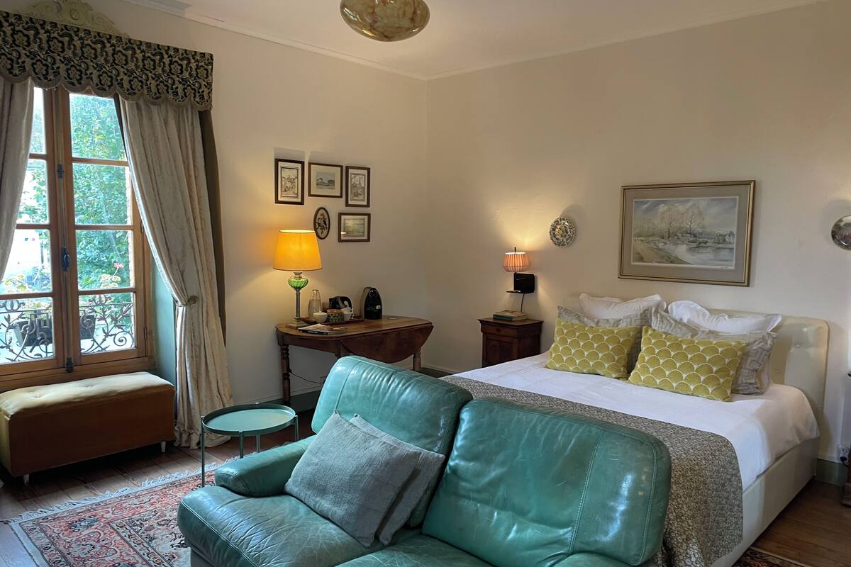 Habitación de hotel con un sofá de cuero verde, una cama con varias almohadas, un escritorio con lámpara y cuadros en la pared.