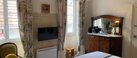 Un dormitorio con cama, cómoda de madera, televisor, silla y ventana con cortinas.