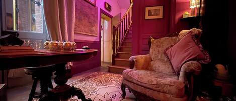 Una habitación con un sillón antiguo, una mesa de madera con objetos decorativos, un tapiz estampado y una escalera con pared roja y cuadros enmarcados.