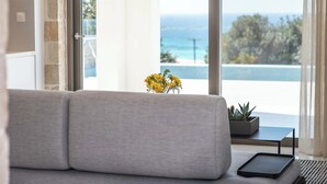 A modern living room with a grey sofa, a small table with a plant, and a view of the sea through large windows. - Actea Seaview Villas | Free* heated pools & 800m sea (Kissamos)