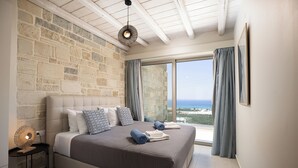 Ein Schlafzimmer mit Steinwand, einem großen Bett mit Kissen, einem Nachttisch mit Lampe und Meerblick durch ein Fenster.