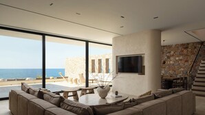 A modern living room with a large sofa, a dining table, and a TV mounted on the wall. - Ormus Villa - An Earthy Beachfront Sanctuary by etouri (Rethymno)
