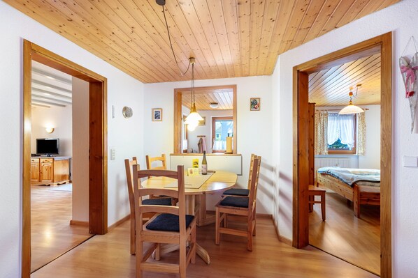 A dining area with wooden chairs and a table, a kitchen with cabinets and a TV, and a bedroom with a bed and window.
