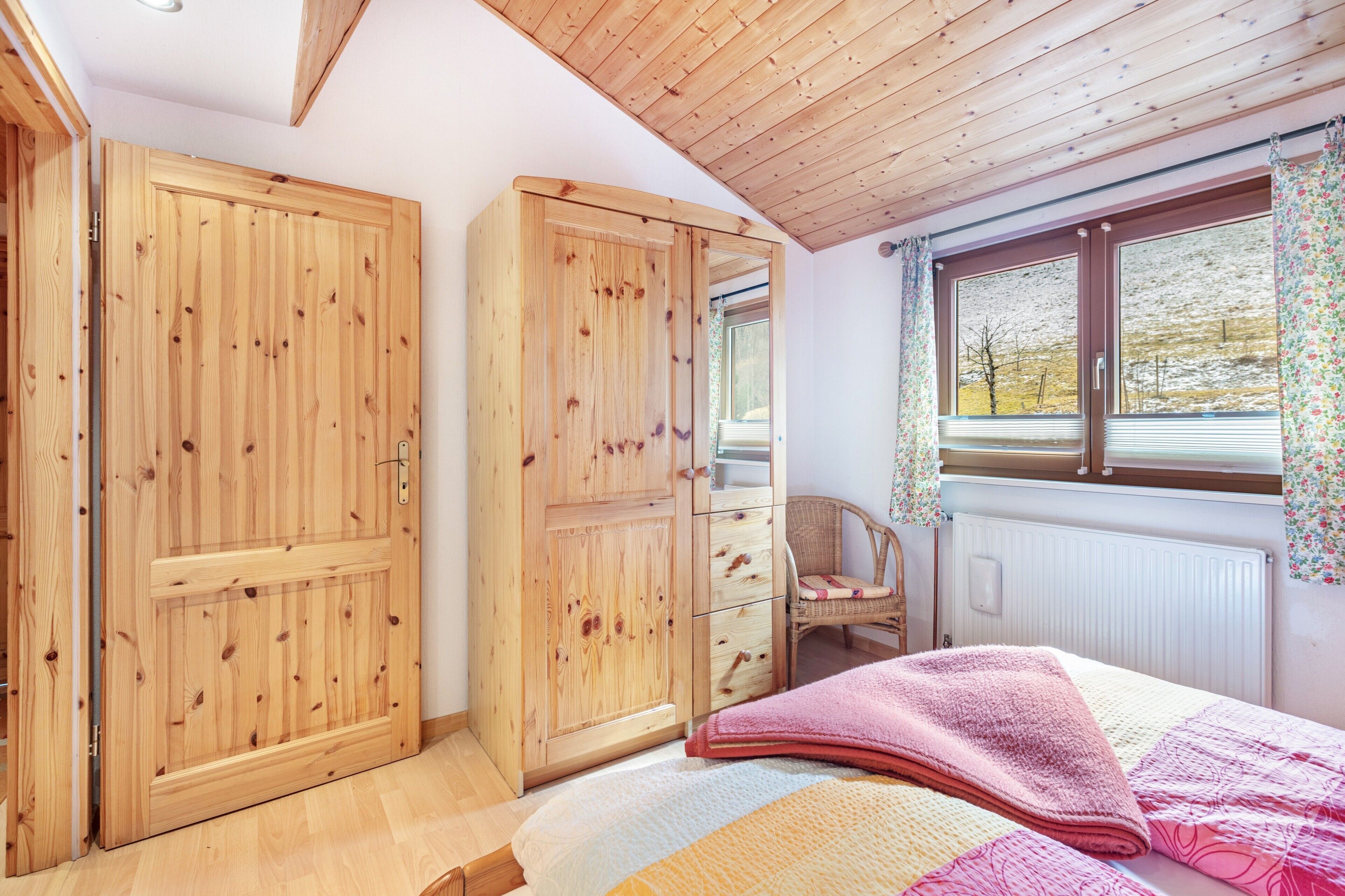 A bedroom with wooden wardrobes, a bed with a pink blanket, a wicker chair, and a window with curtains.