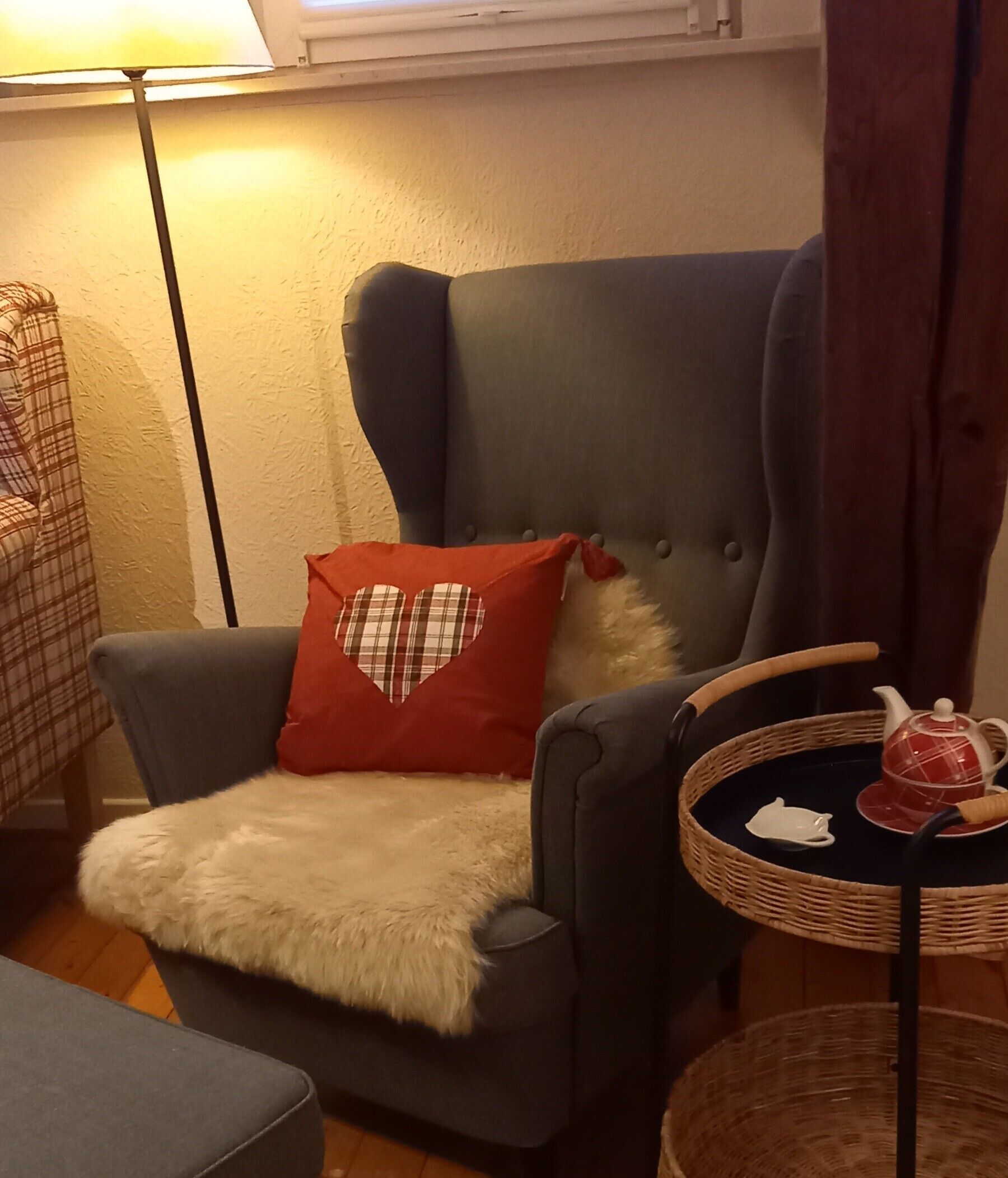 Una habitación de esquina con un sillón gris, un cojín rojo con forma de corazón, una mesita redonda con una tetera y una cesta de mimbre.