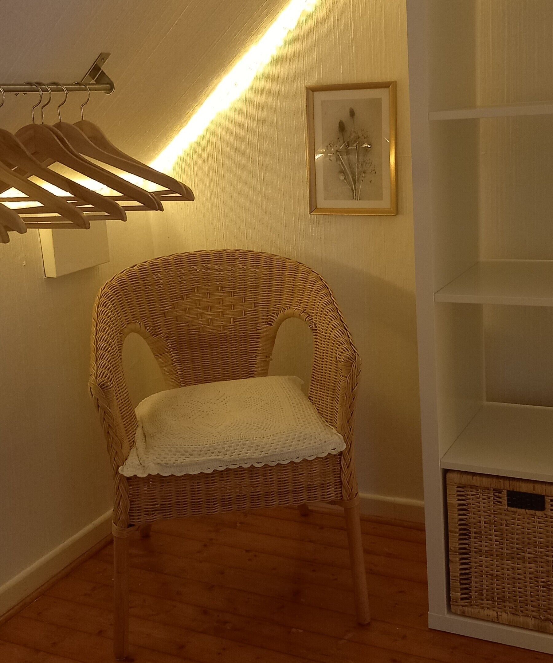 Une chaise en osier avec un coussin blanc, un luminaire fixé au mur, un tableau encadré et une étagère blanche.