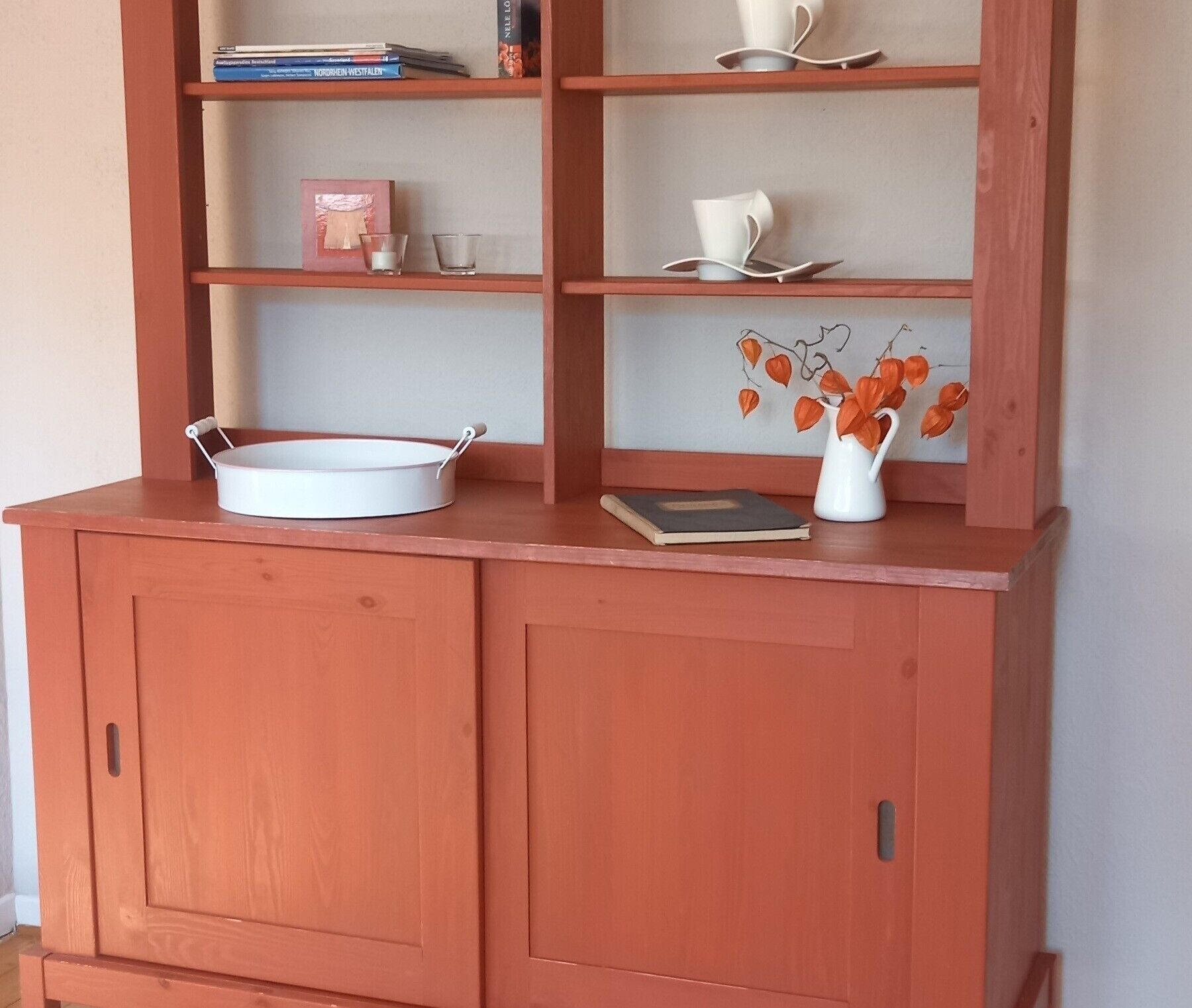 Ein Holzschrank mit Regalen, ein weißer Topf, ein Buch und eine Vase mit Blättern.