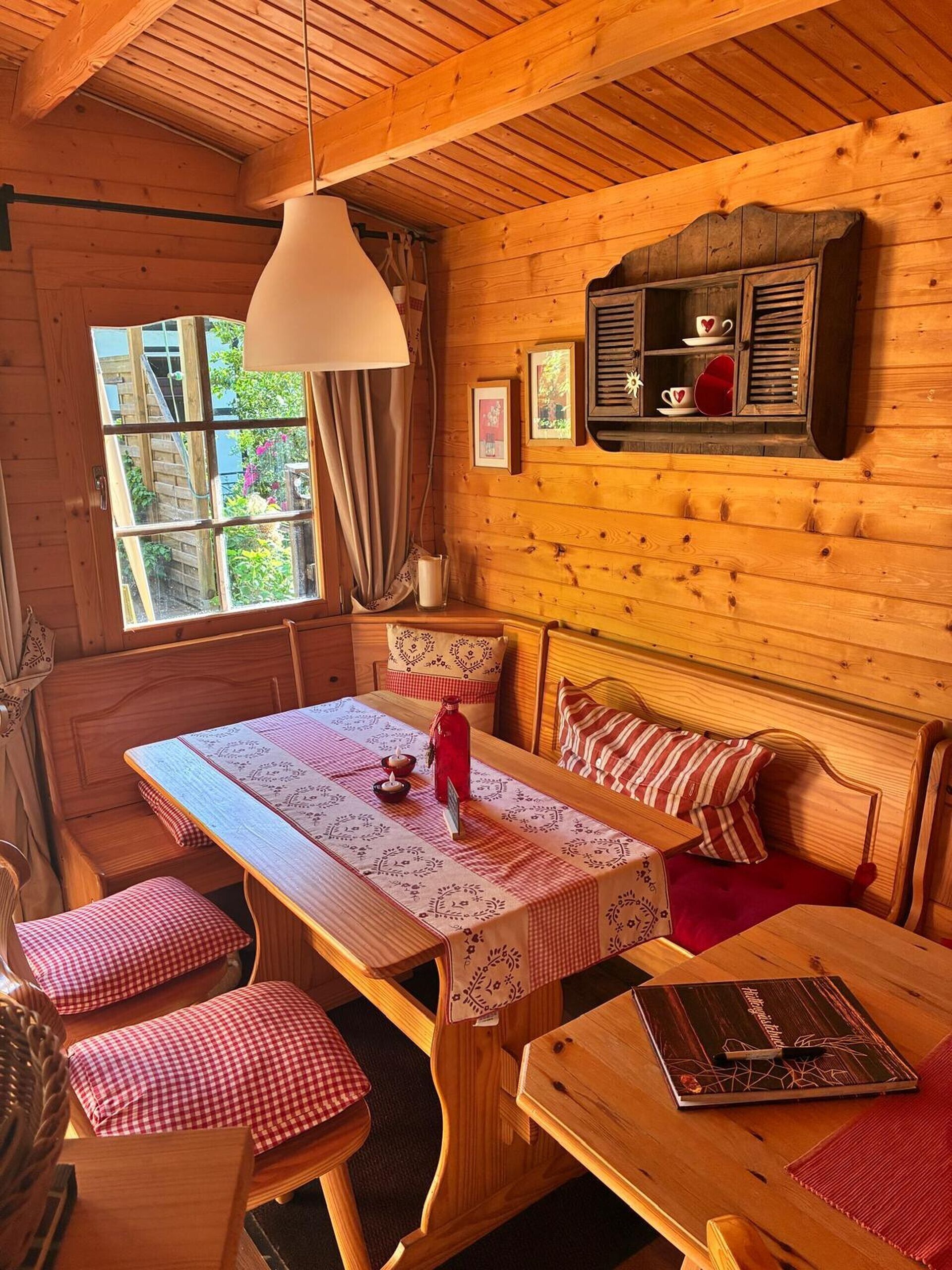 Ein gemütliches Blockhaus-Interieur mit einem Holzesstisch, Bänken, einer Bank mit Sitzkissen, einem an der Wand befestigten Regal mit Dekorationen und einem Fenster mit Vorhängen.