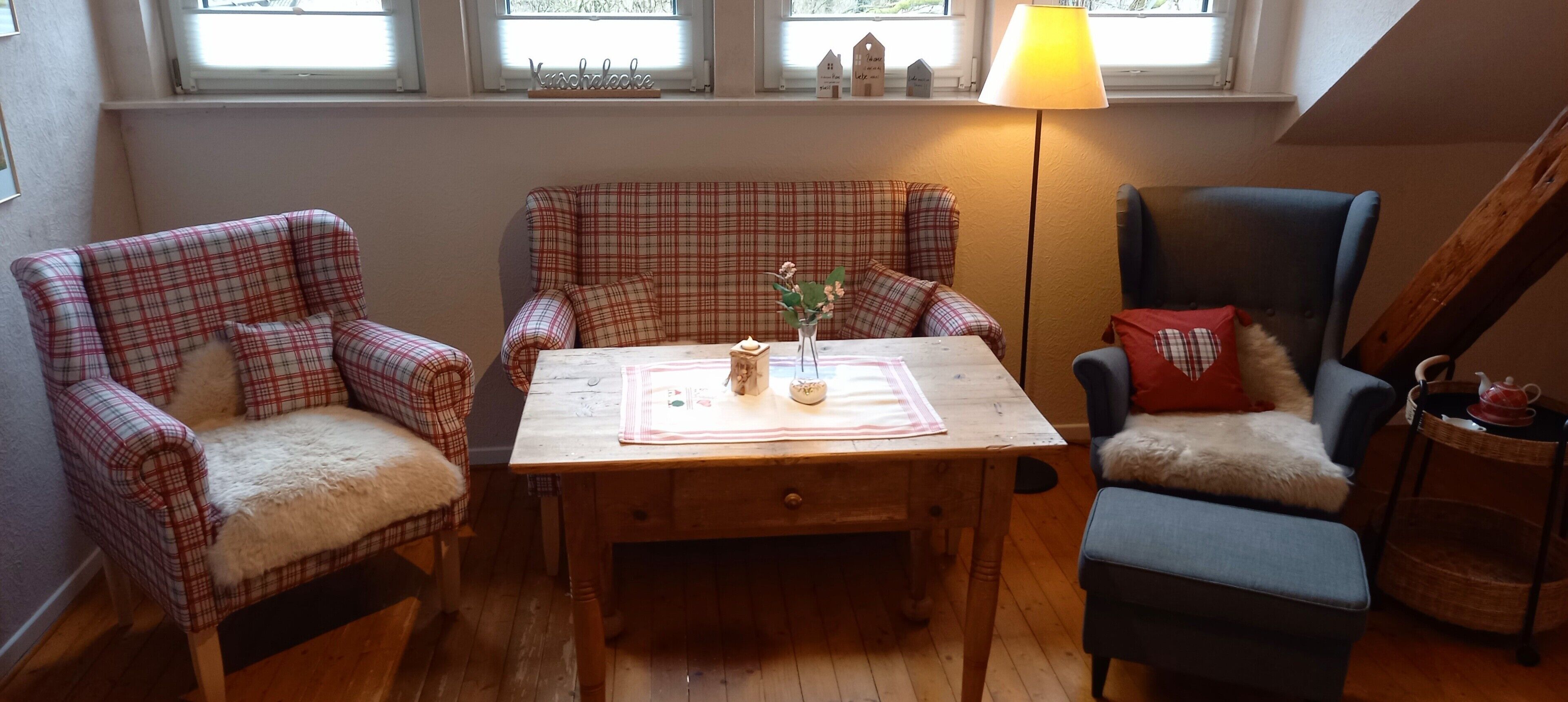 Ein gemütliches Zimmer mit einem Holztisch, Karo-Stühlen, einer Lampe und Blick ins Freie durch ein Fenster.