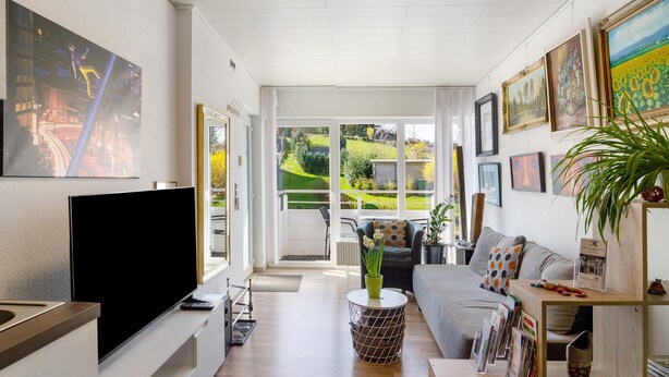 A modern living room with a flat-screen TV, a sofa, a coffee table, and a view of a garden through large windows.