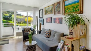 A living room with a sofa, a coffee table, and a bookshelf. There are framed pictures on the wall and a plant on the bookshelf.