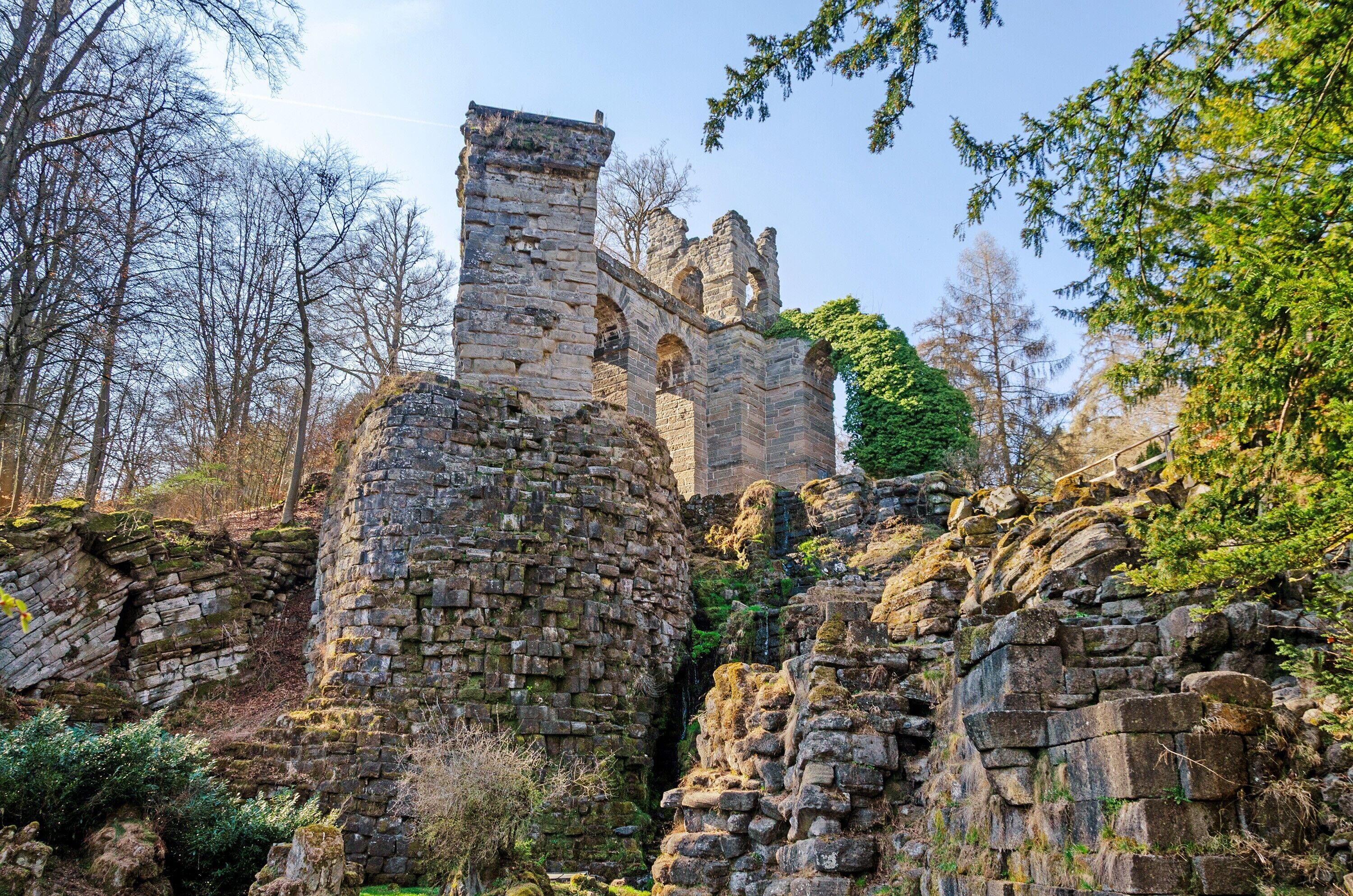 Eine historische Steinruine mit Bogenfenstern und einem Turm, umgeben von Bäumen und Vegetation.